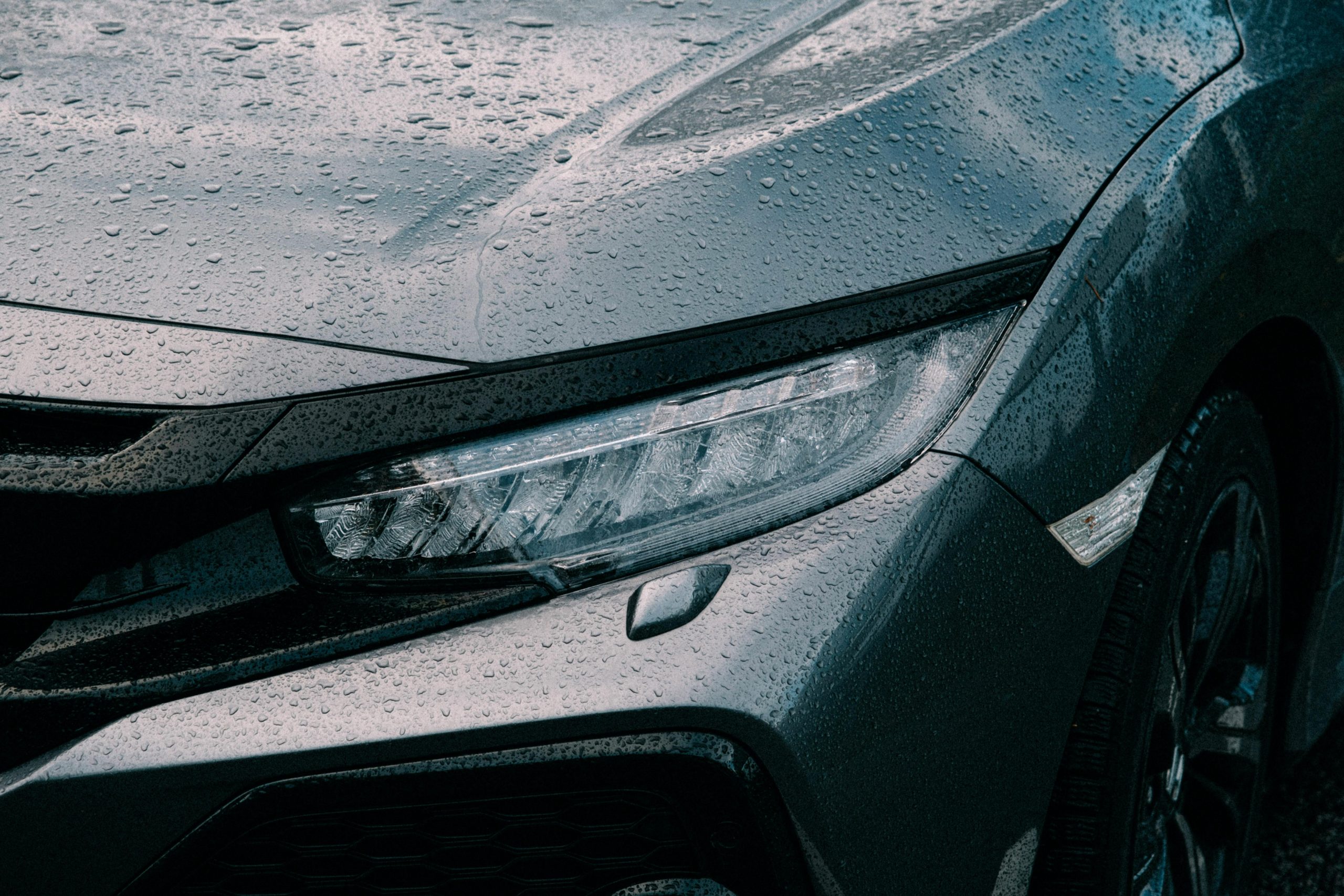 Detailed view of a car's headlight and hood with water drops, reflecting a rainy day.