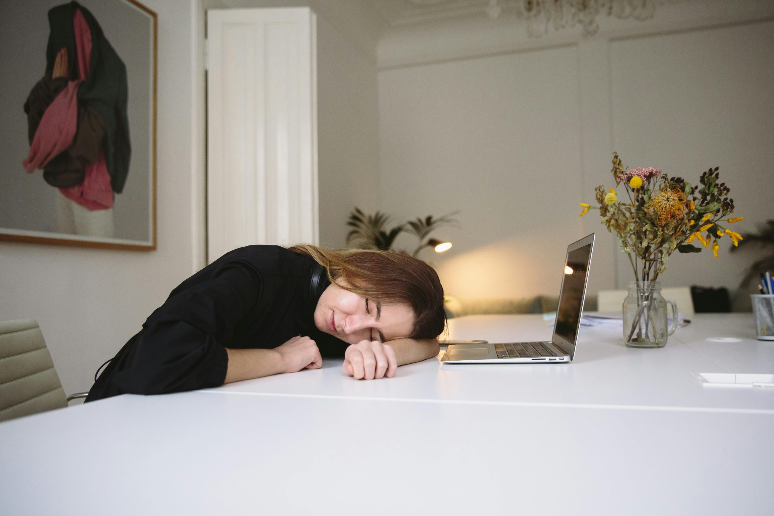 Exhausted woman sleeping at a desk next to a laptop, surrounded by flower arrangements in an office environment.