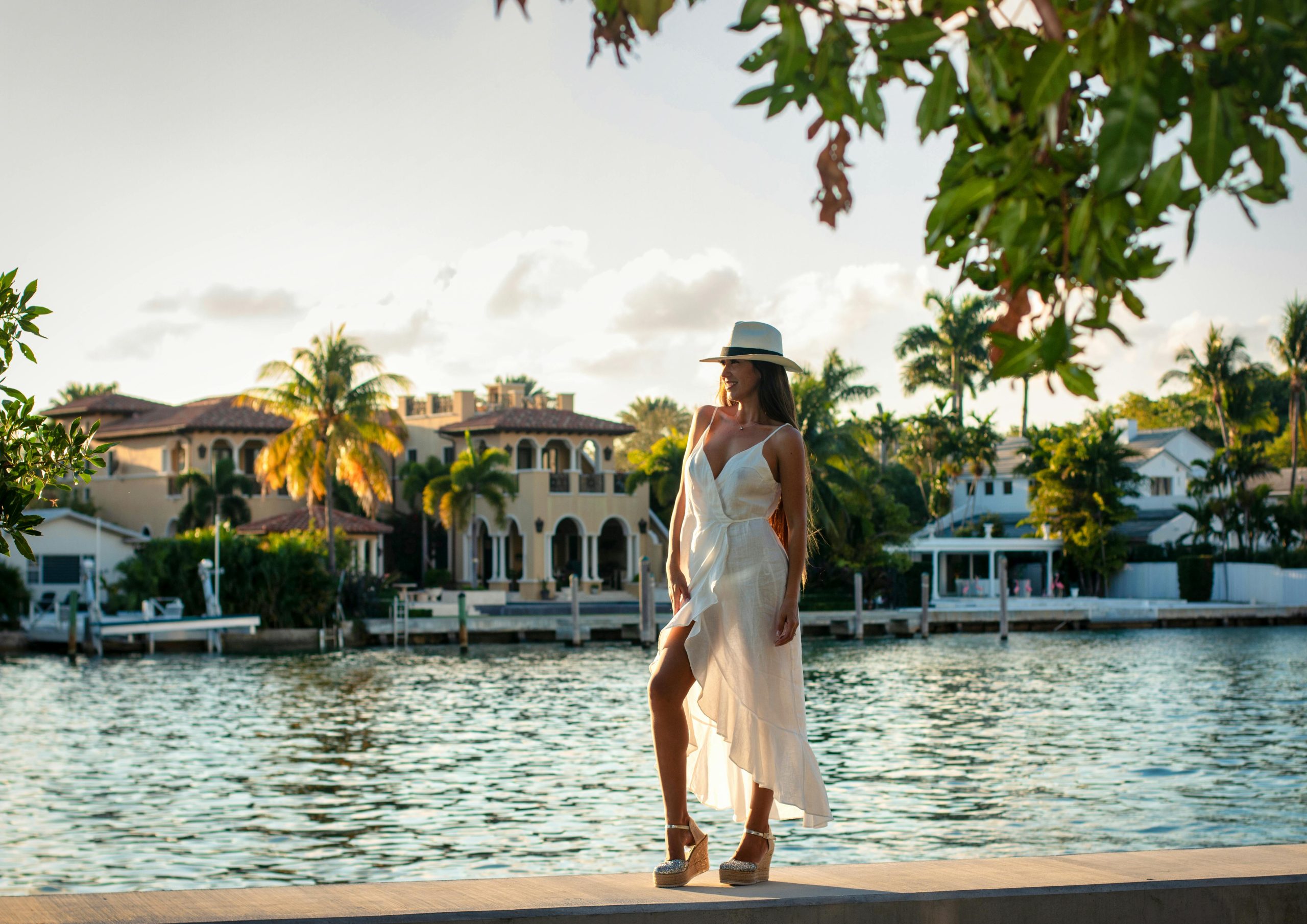 Chic woman in white dress and hat posing by scenic Miami Beach waterfront at sunset.