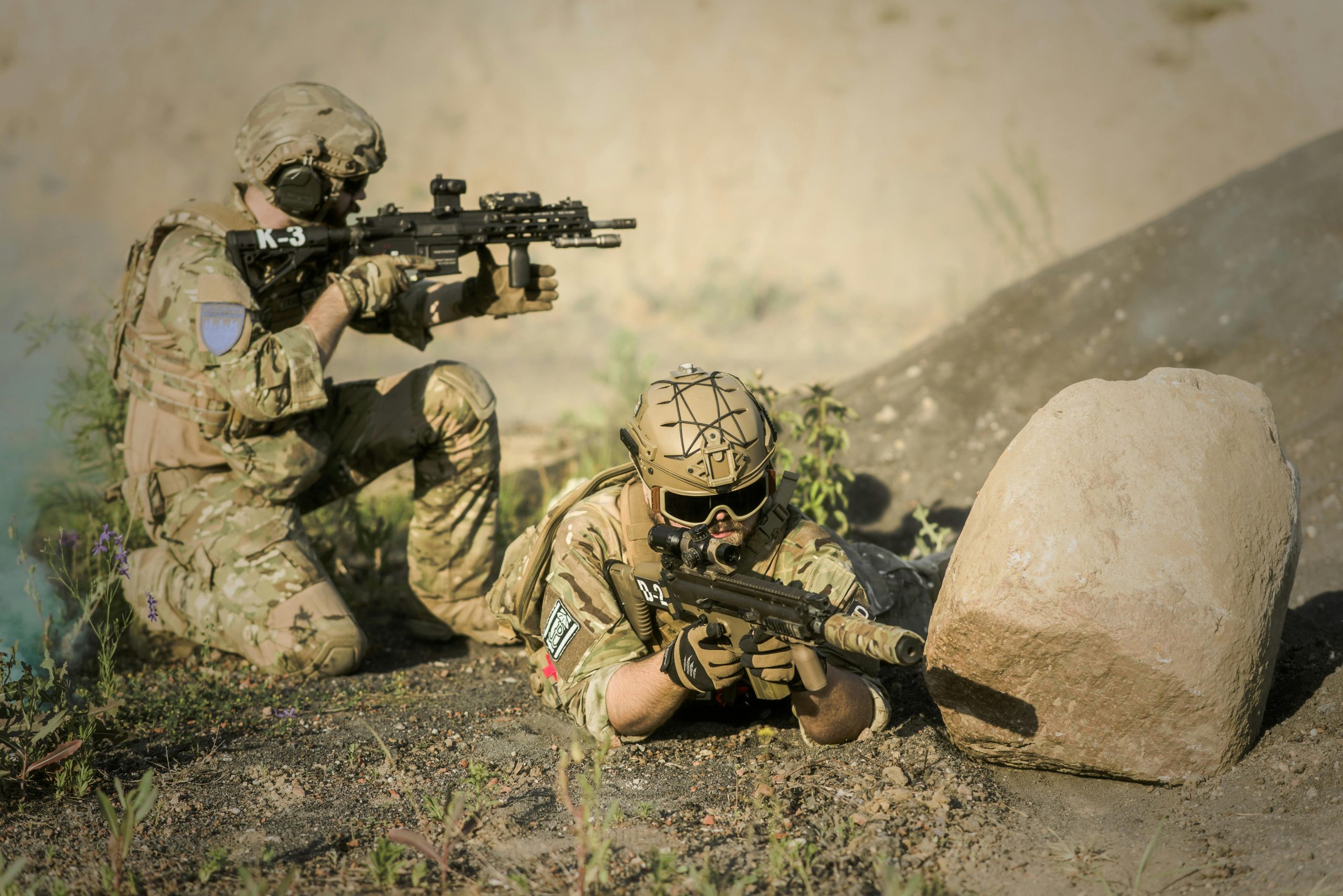 Two soldiers in camouflage uniforms practice tactical maneuvers in a desert environment, exhibiting teamwork and precision.