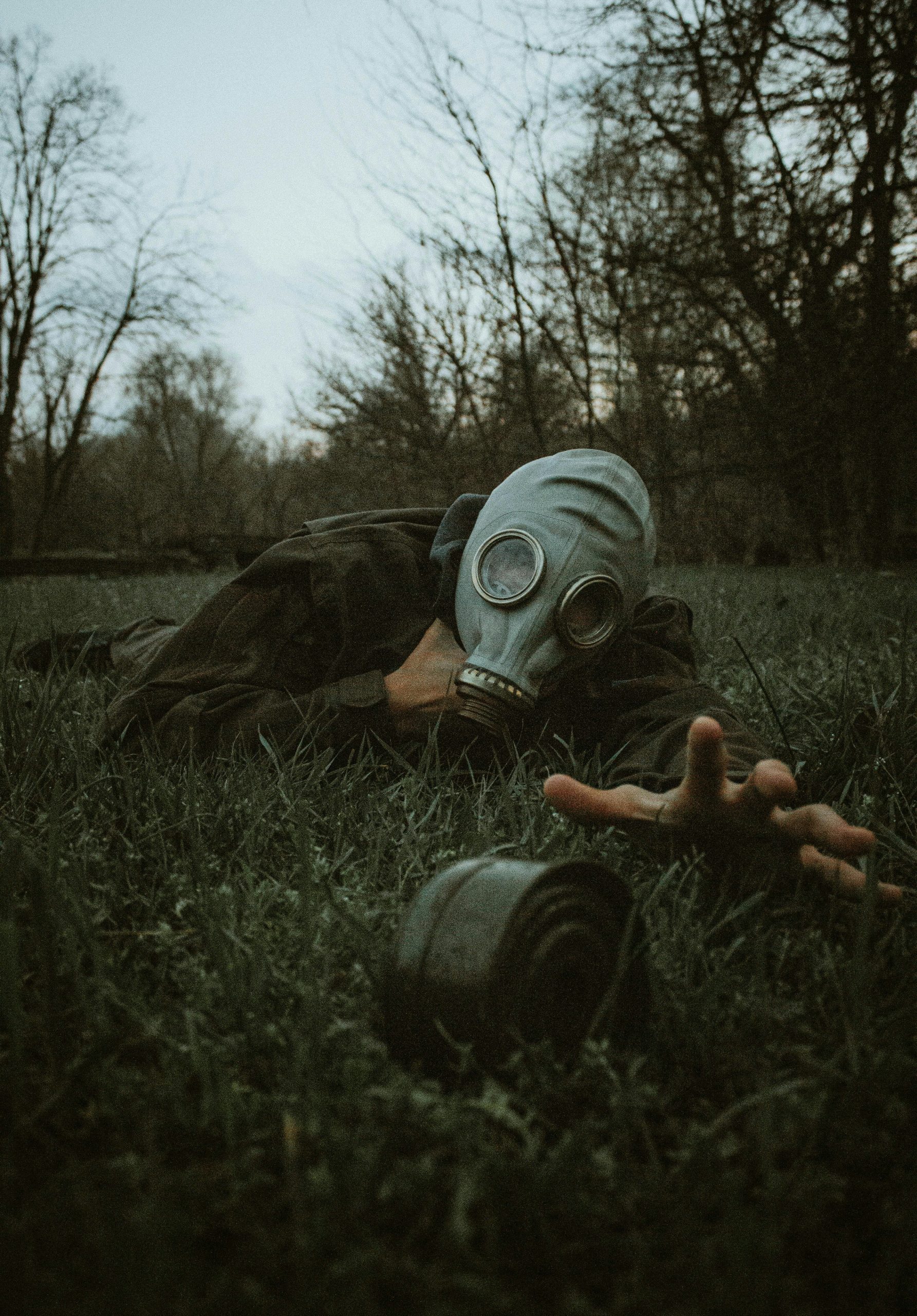 A person wearing a gas mask reaches across the grass in a moody forest setting.