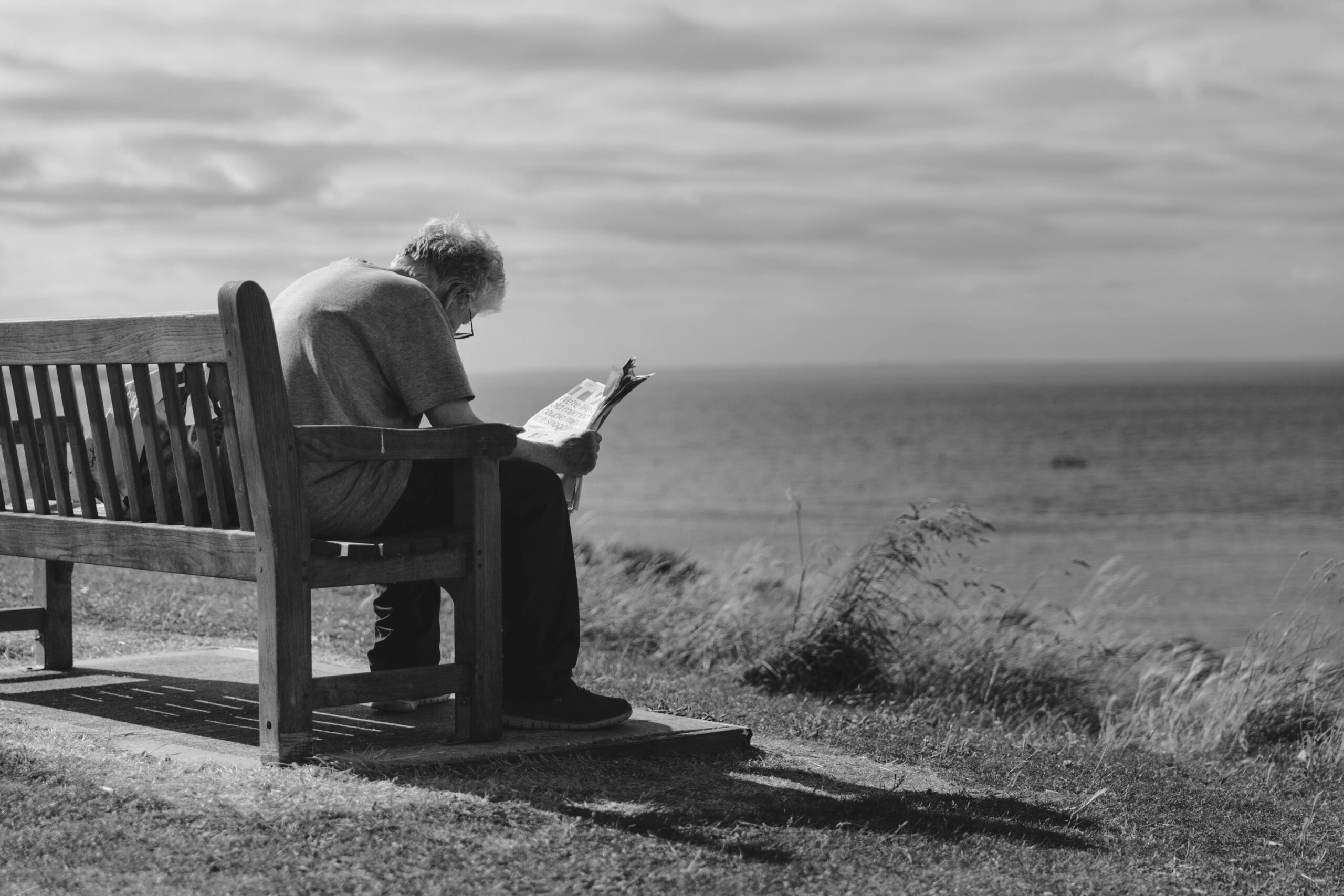 À quel âge sommes-nous le plus heureux ? Une réponse (presque) contre-intuitive 4 A contemplative scene of an elderly man reading a newspaper on a seaside bench with cloudy skies.