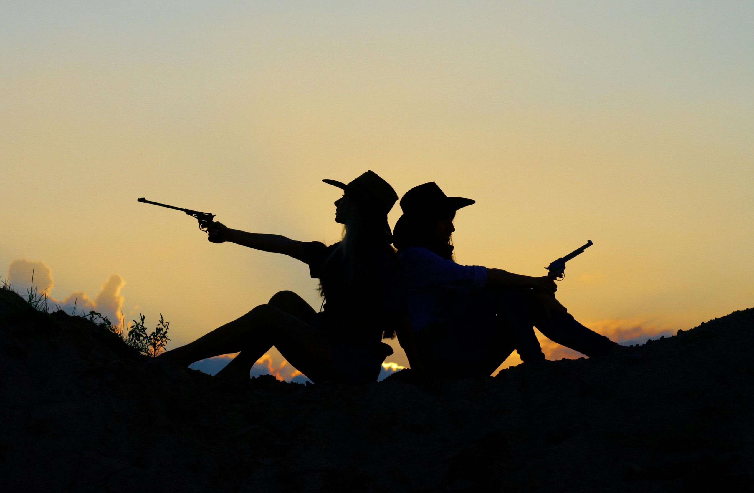 Silhouette of cowgirls posing with guns against a dramatic sunset sky, evoking a western theme.