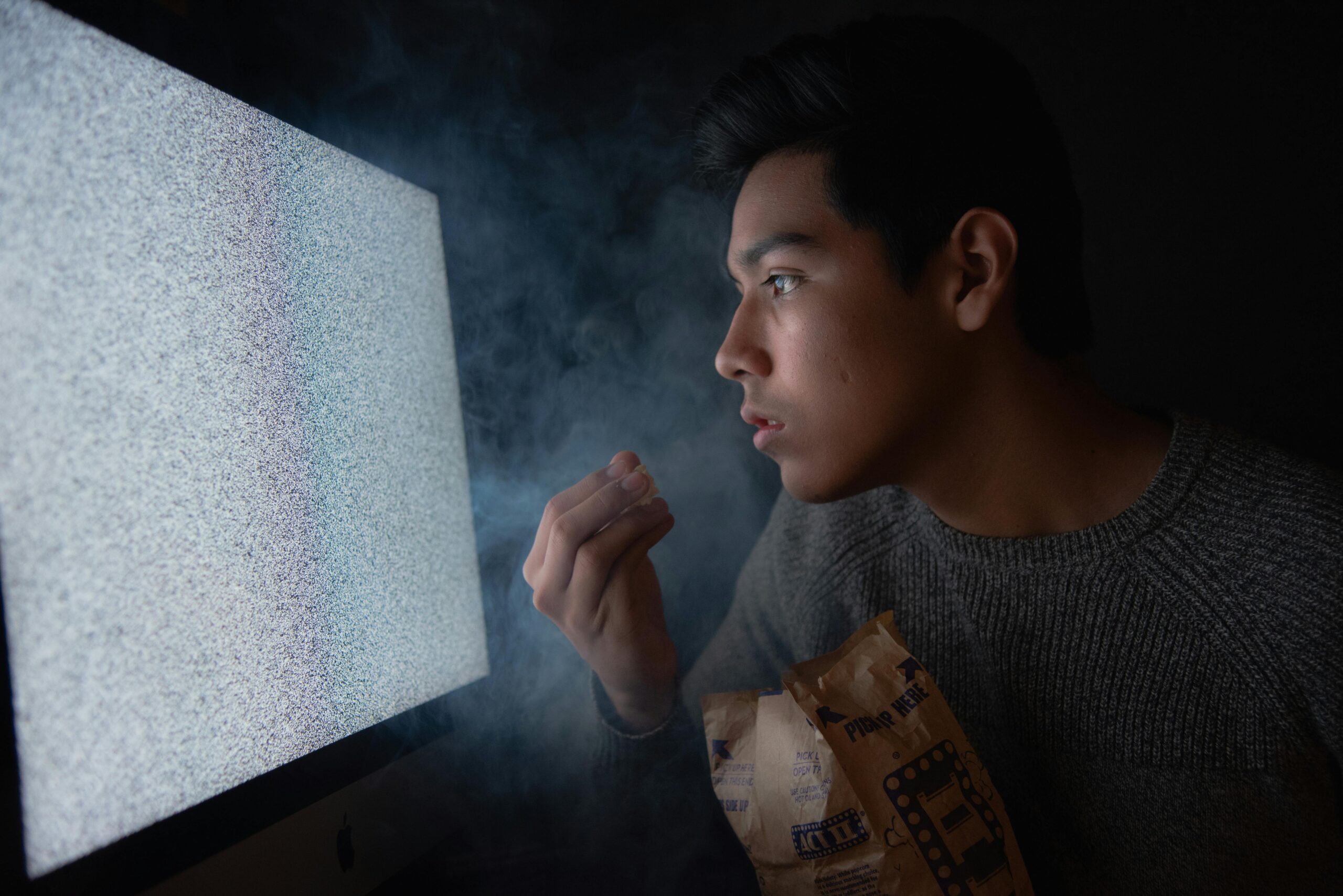 A young man engaged in watching TV static with popcorn in a dark room, creating a mysterious atmosphere.
