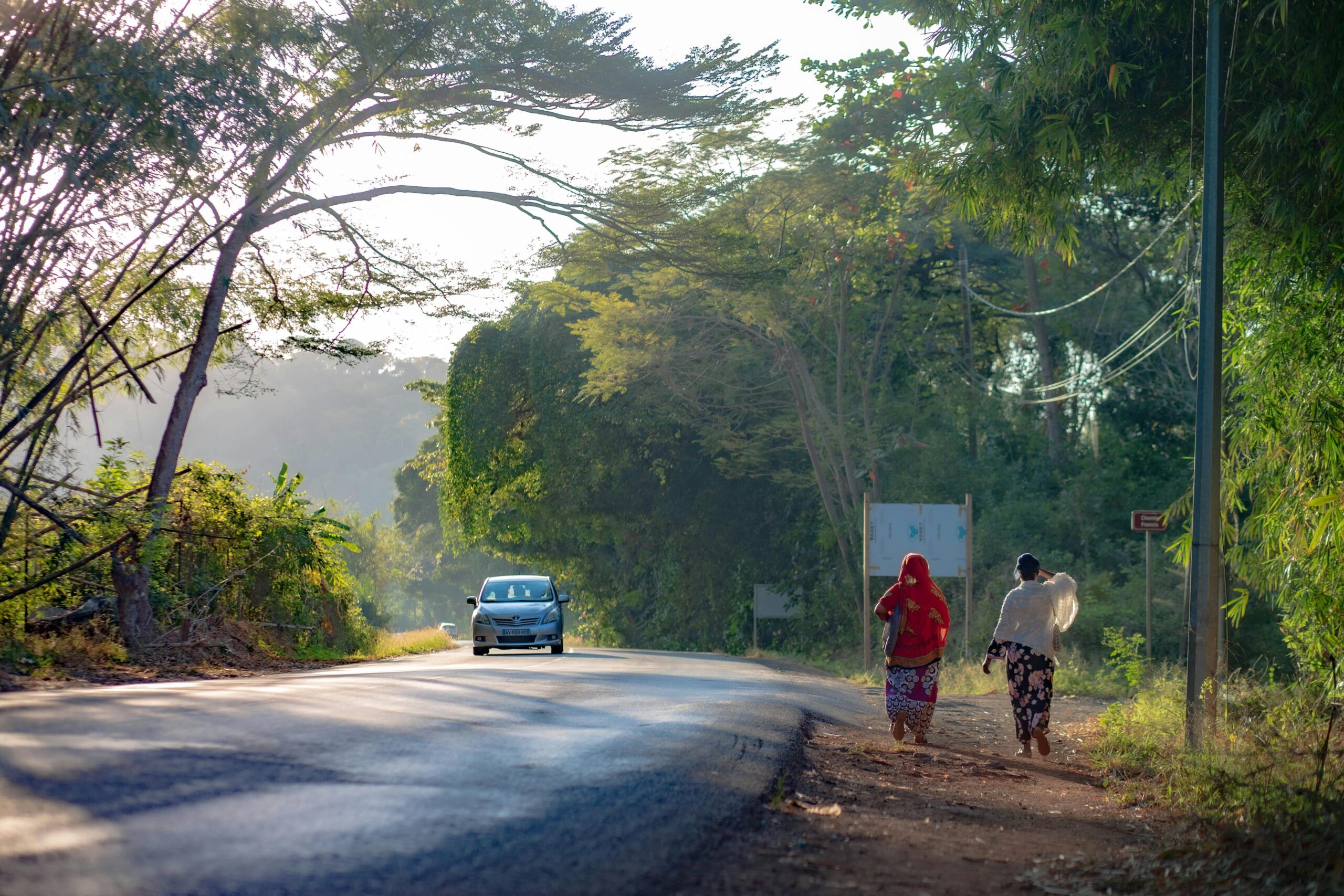 A serene road in Dembéni, Mayotte, with two pedestrians walking along lush greenery.