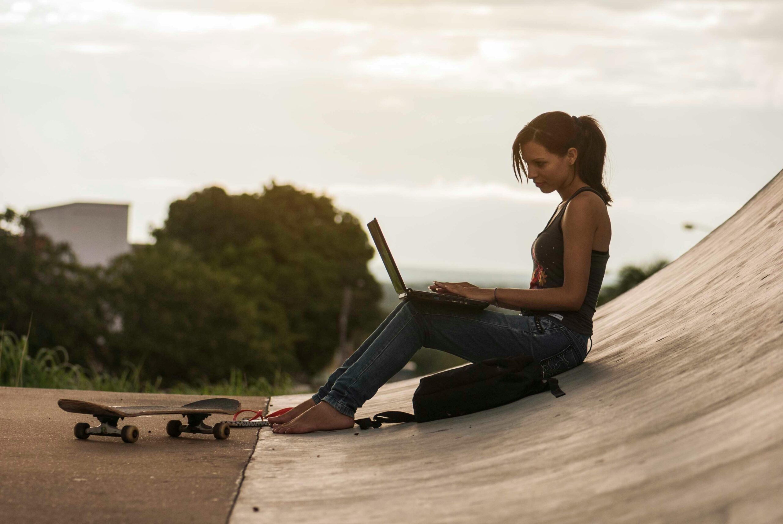 A young woman using a laptop while sitting at a skate park during sunset.