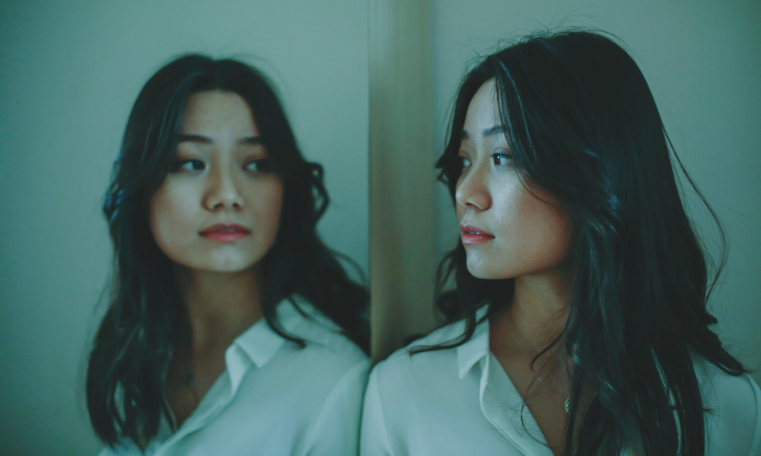 A thoughtful young woman gazes at her reflection in a serene indoor setting.