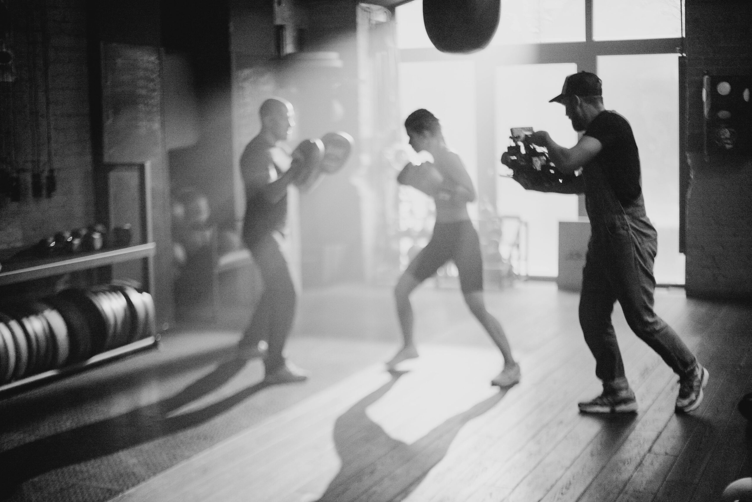 Black and white image capturing a boxing training session with a cameraman filming.