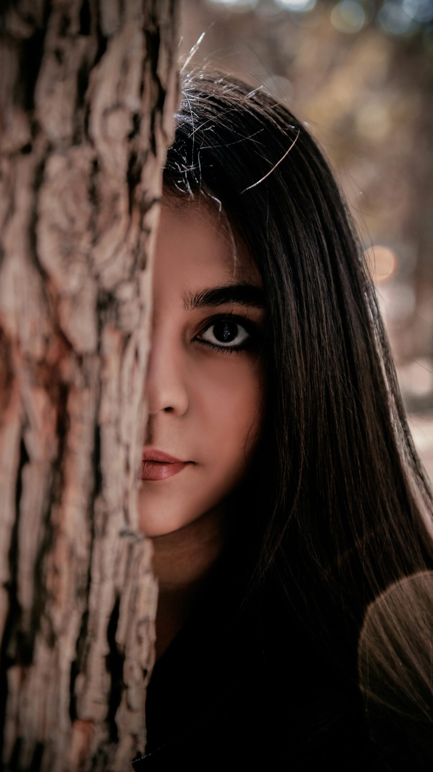 Close-up of a woman partially hidden behind a tree, conveying a sense of mystery.