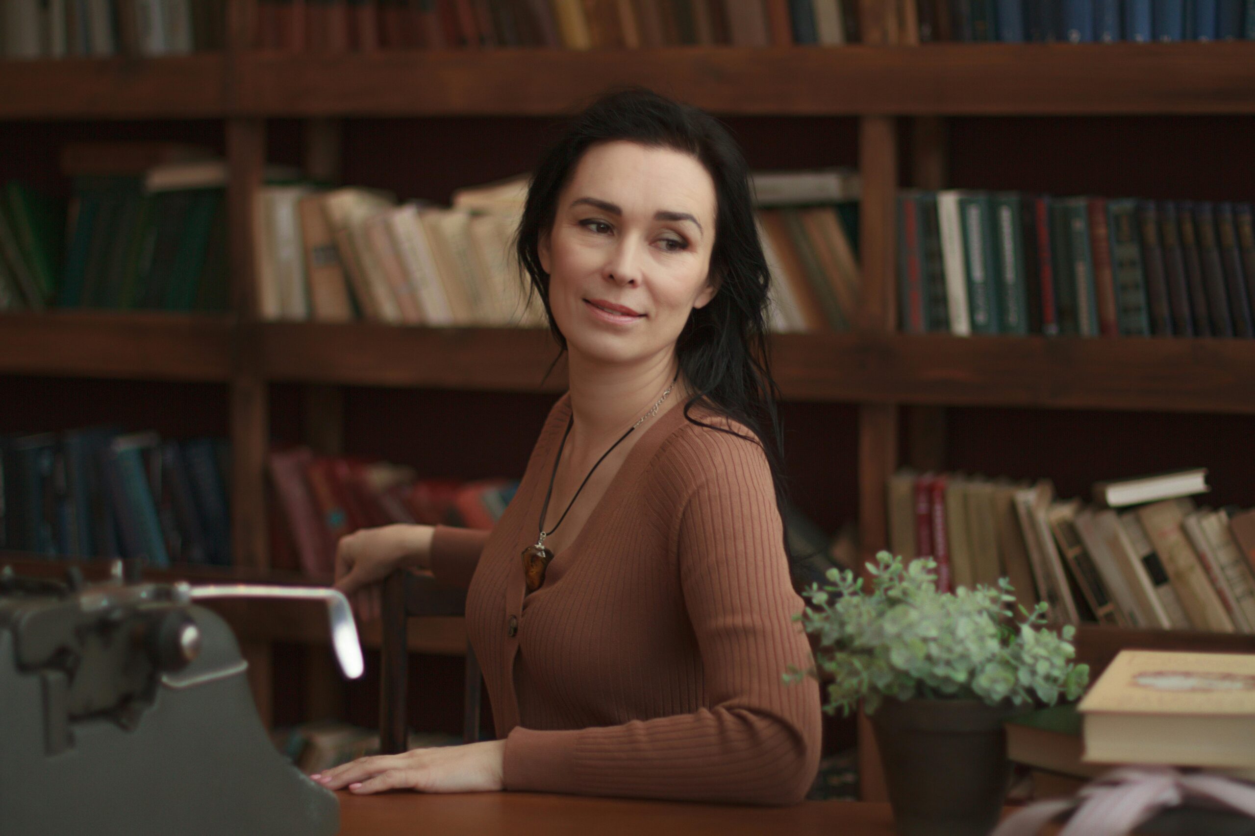 A woman in a brown outfit sitting in a cozy library setting with a typewriter.