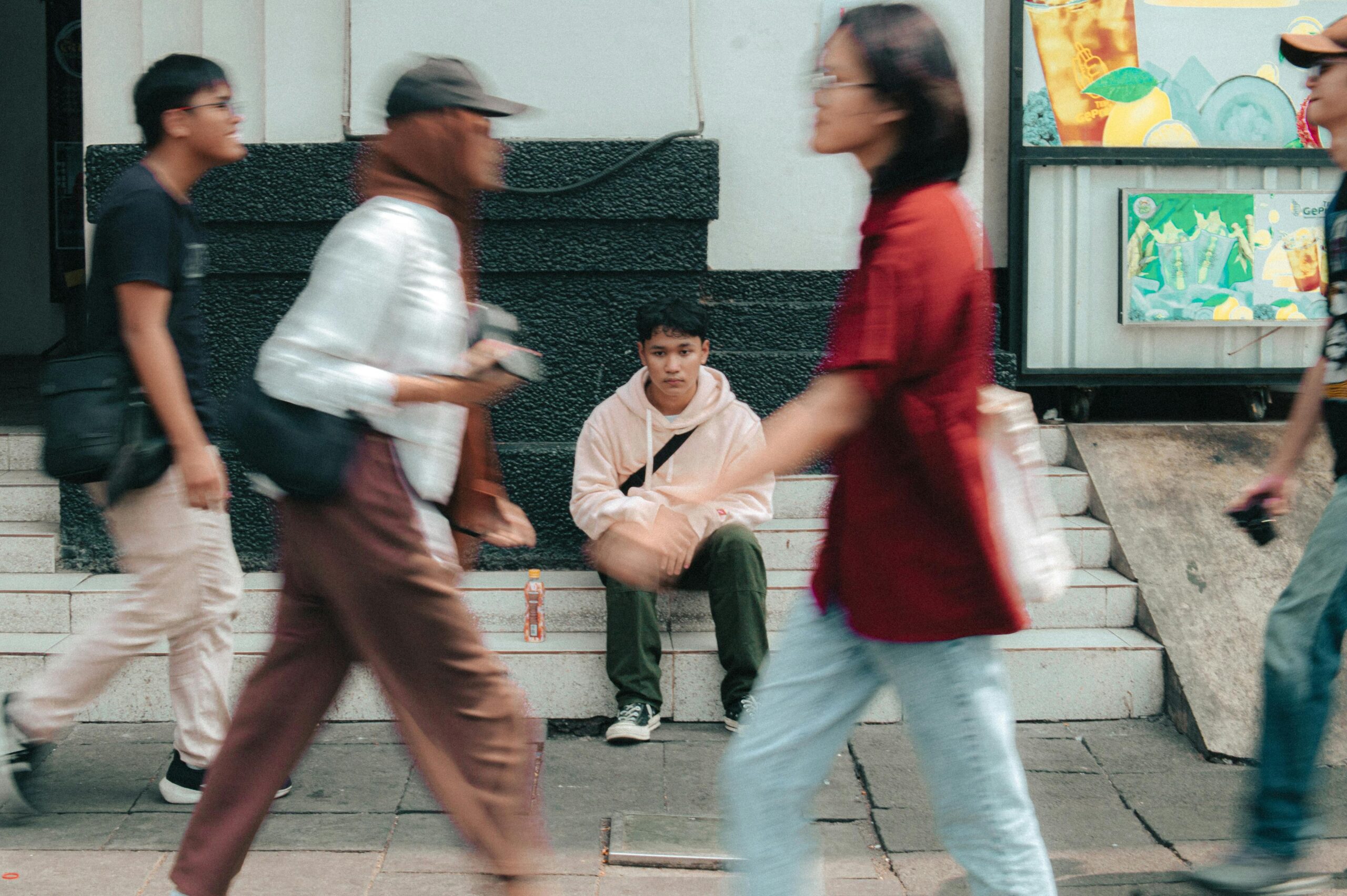 A young man in a pink hoodie sitting on steps as people walk by in motion blur.