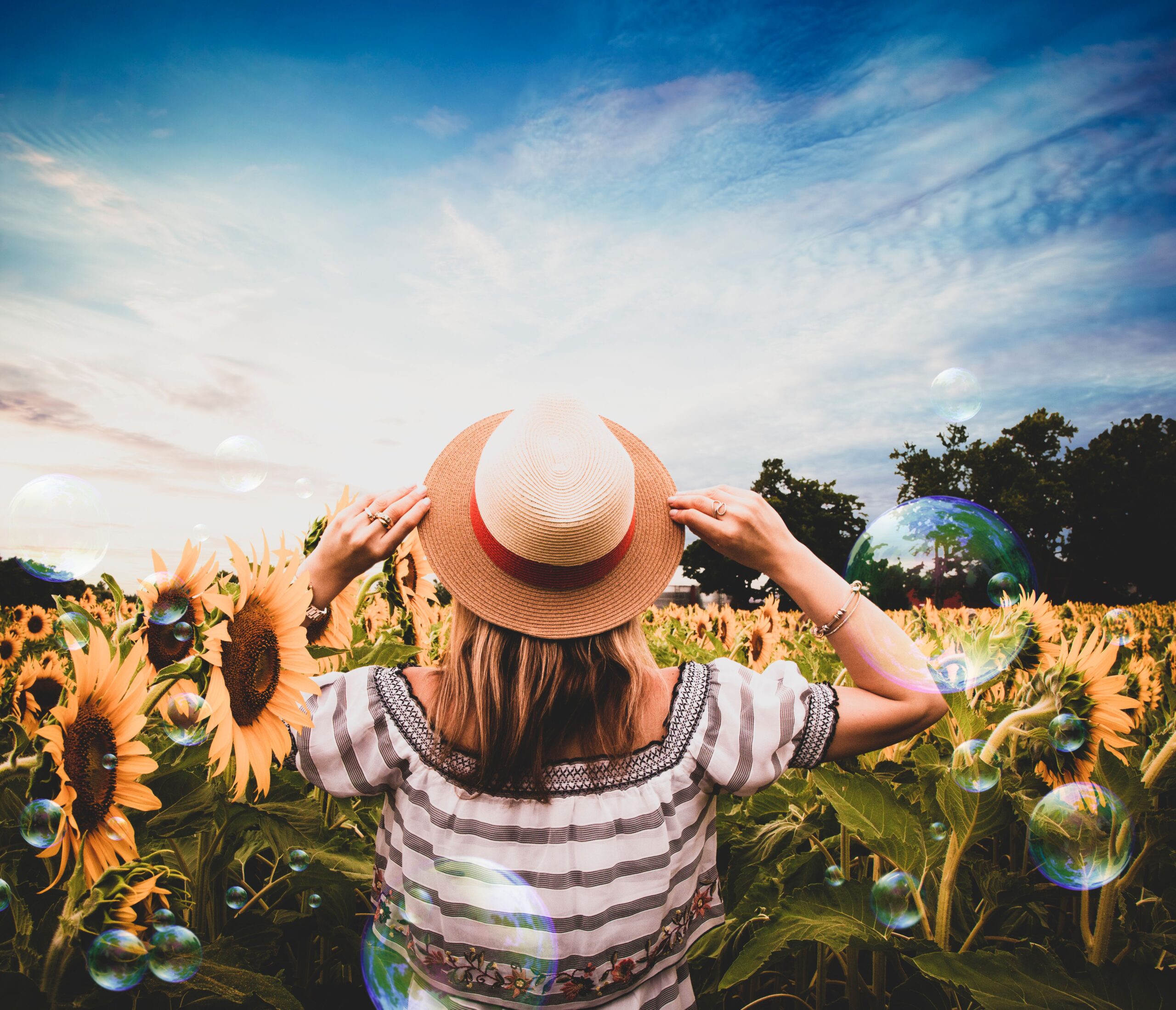 A woman stands in a vibrant sunflower field, enjoying the summer day with bubbles floating around.