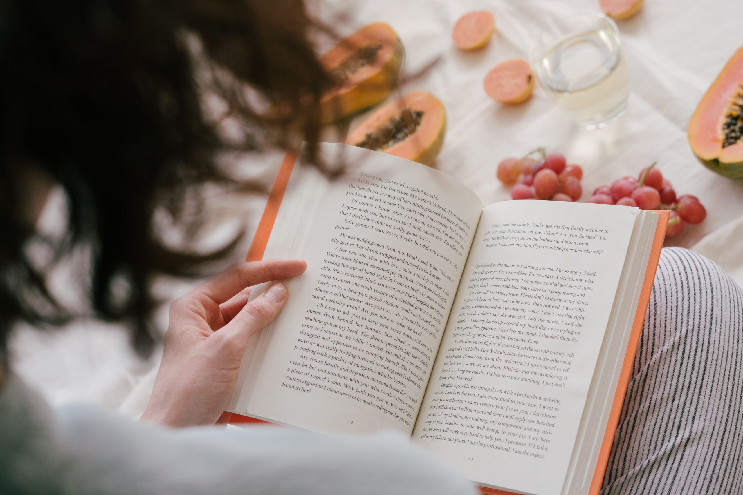 30 livres à lire pour apprendre à écrire comme un pro 3 A person enjoys reading a book surrounded by fresh papaya, grapes, and a drink, creating a cozy indoor scene.