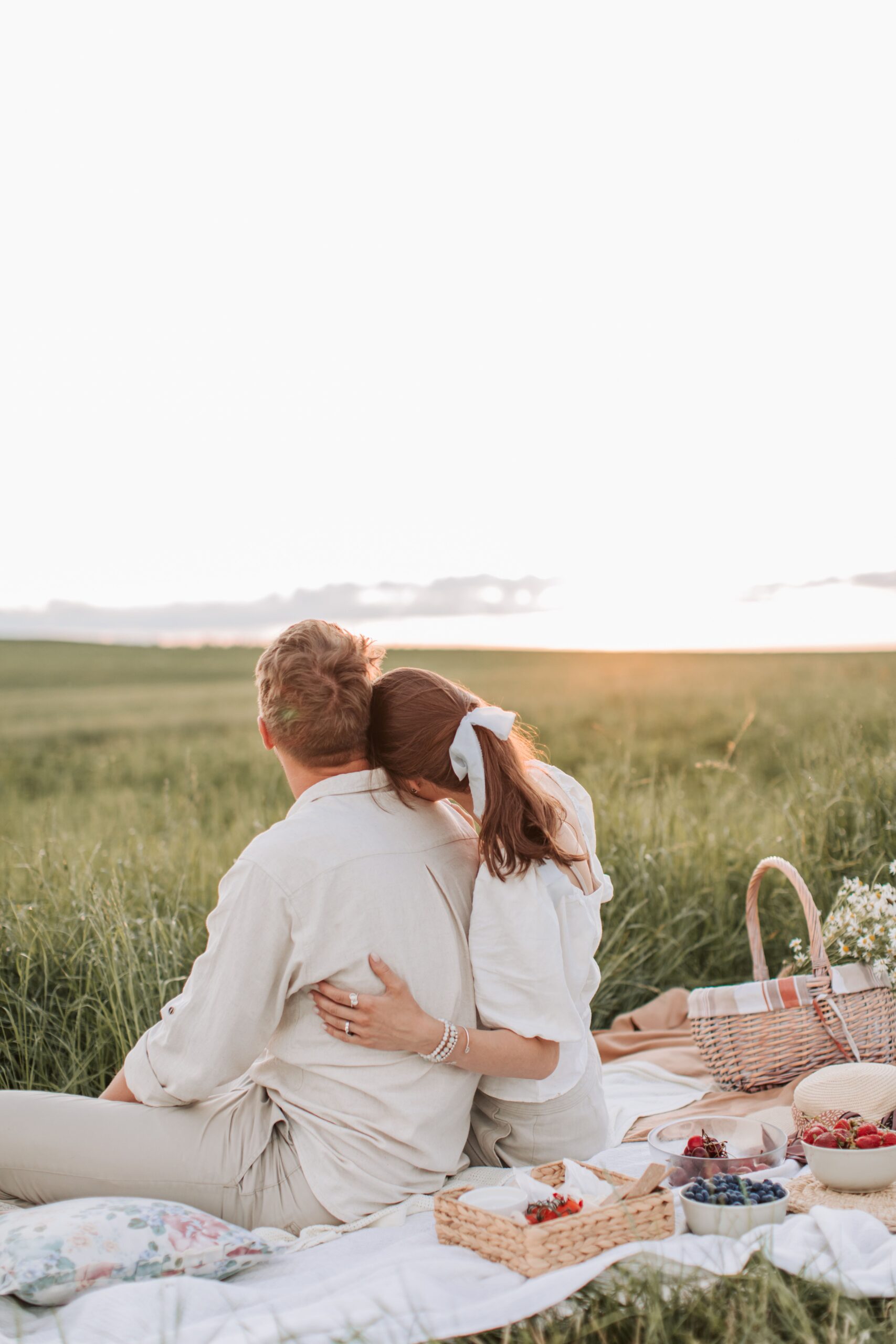 Couple enjoying a romantic sunset picnic in a grassy field with a variety of fruits and a cozy blanket.