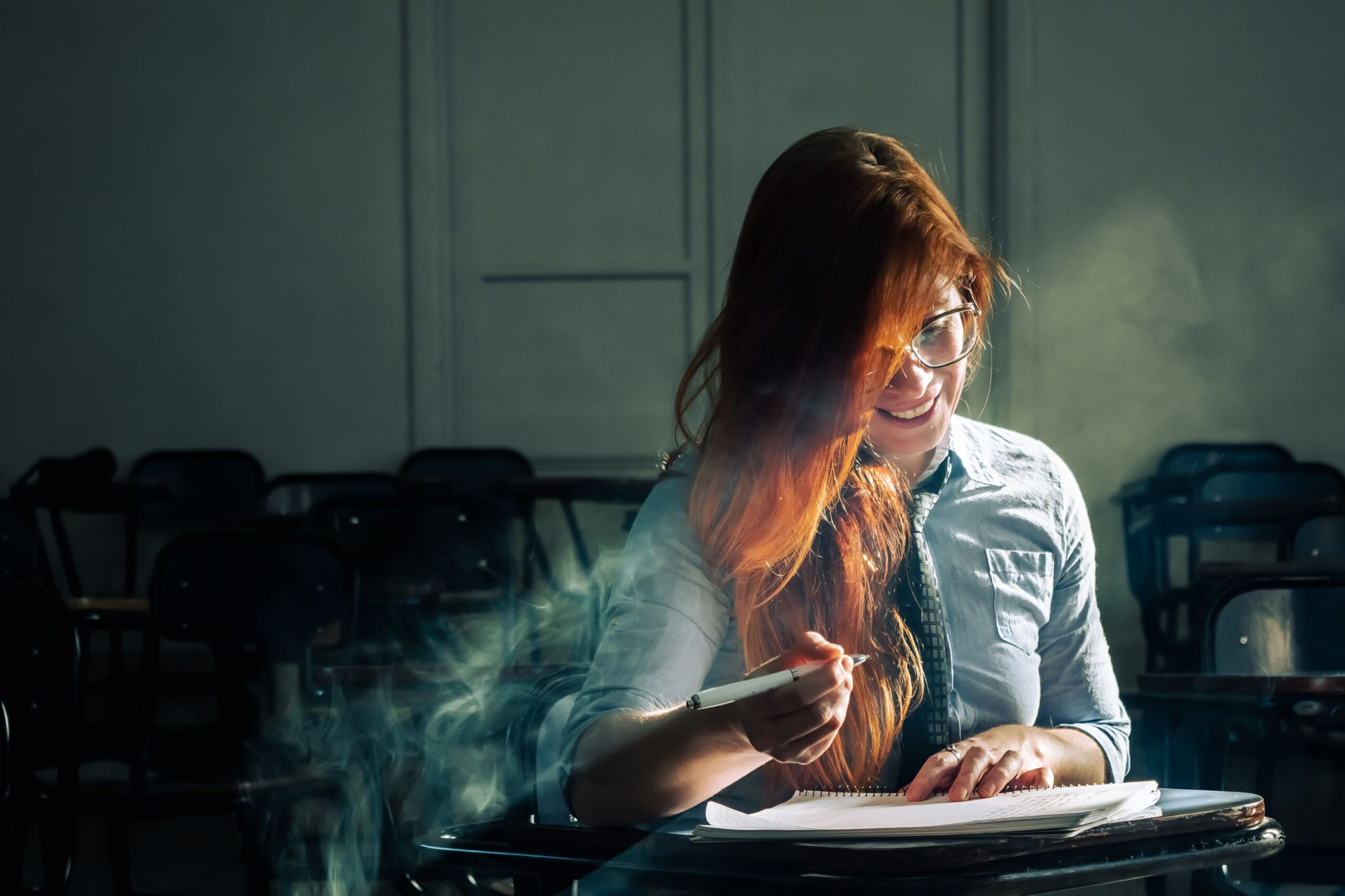 A young woman sits alone in a dimly lit classroom, writing and studying intently.